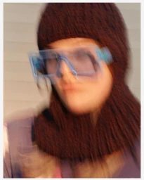 Portrait of a person wearing a brown knitted balaclava and light blue oversized glasses with motion blur.