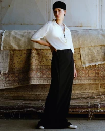 Model wearing a white polo shirt, black skirt, and head covering standing against rolled rugs by photographer Osman Ozel.