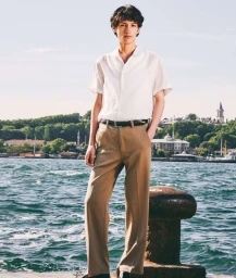 Young man in a white short-sleeve shirt and khaki trousers standing by the water with a city shoreline in the background.