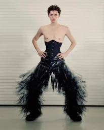 Man wearing a black corset and fringed pants standing against a slatted background