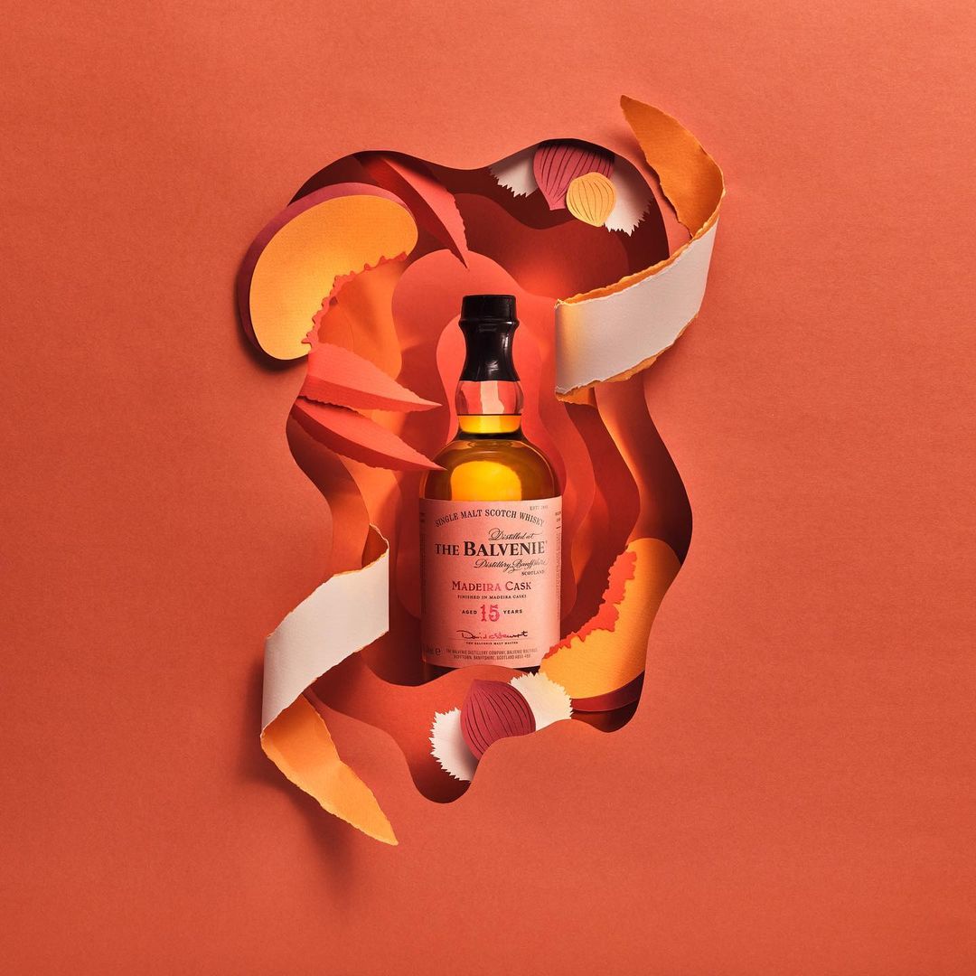 Studio shot of Balvenie 15 Year Madeira Cask single malt Scotch whisky bottle against coral backdrop with paper art design.