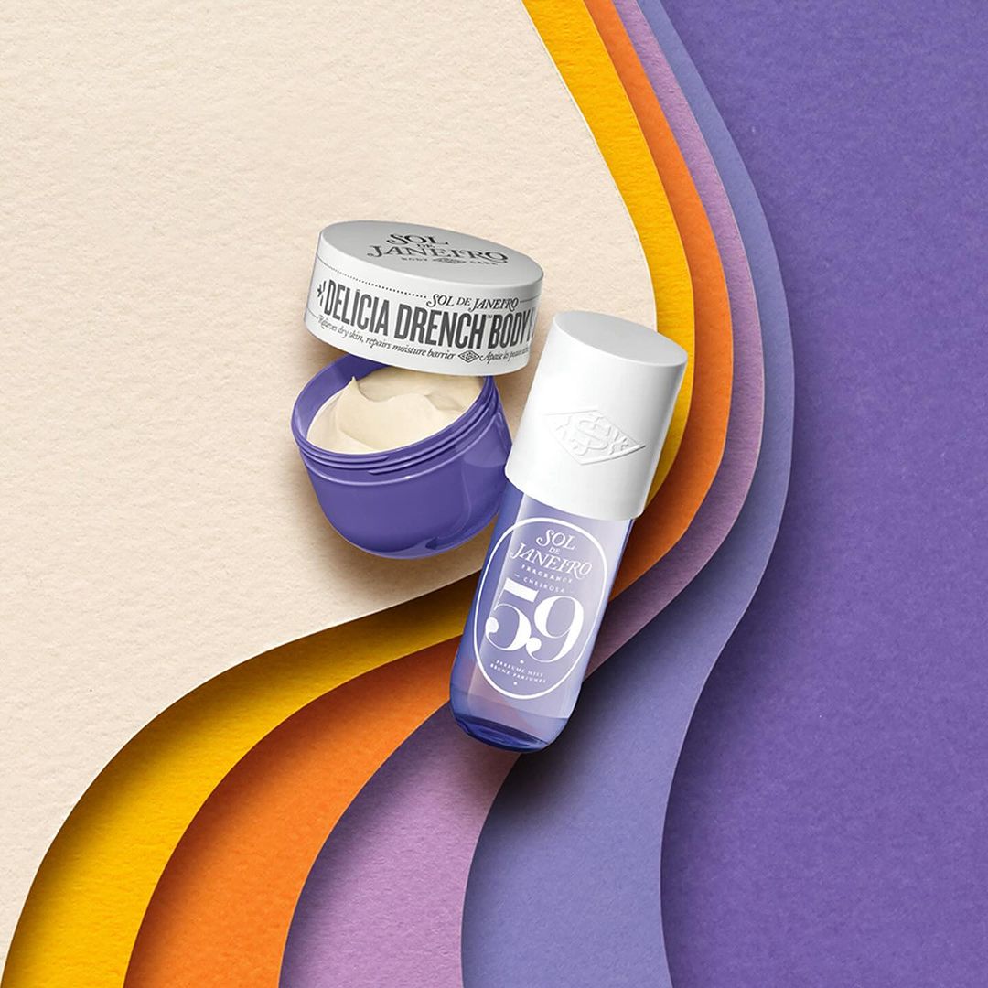 Still life shot of Sol de Janeiro body creams and fragrance mist on textured paper layers in yellow, orange, and purple tones.