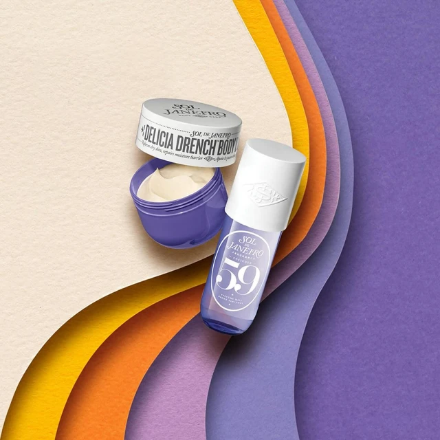 Still life shot of Sol de Janeiro body creams and fragrance mist on textured paper layers in yellow, orange, and purple tones.