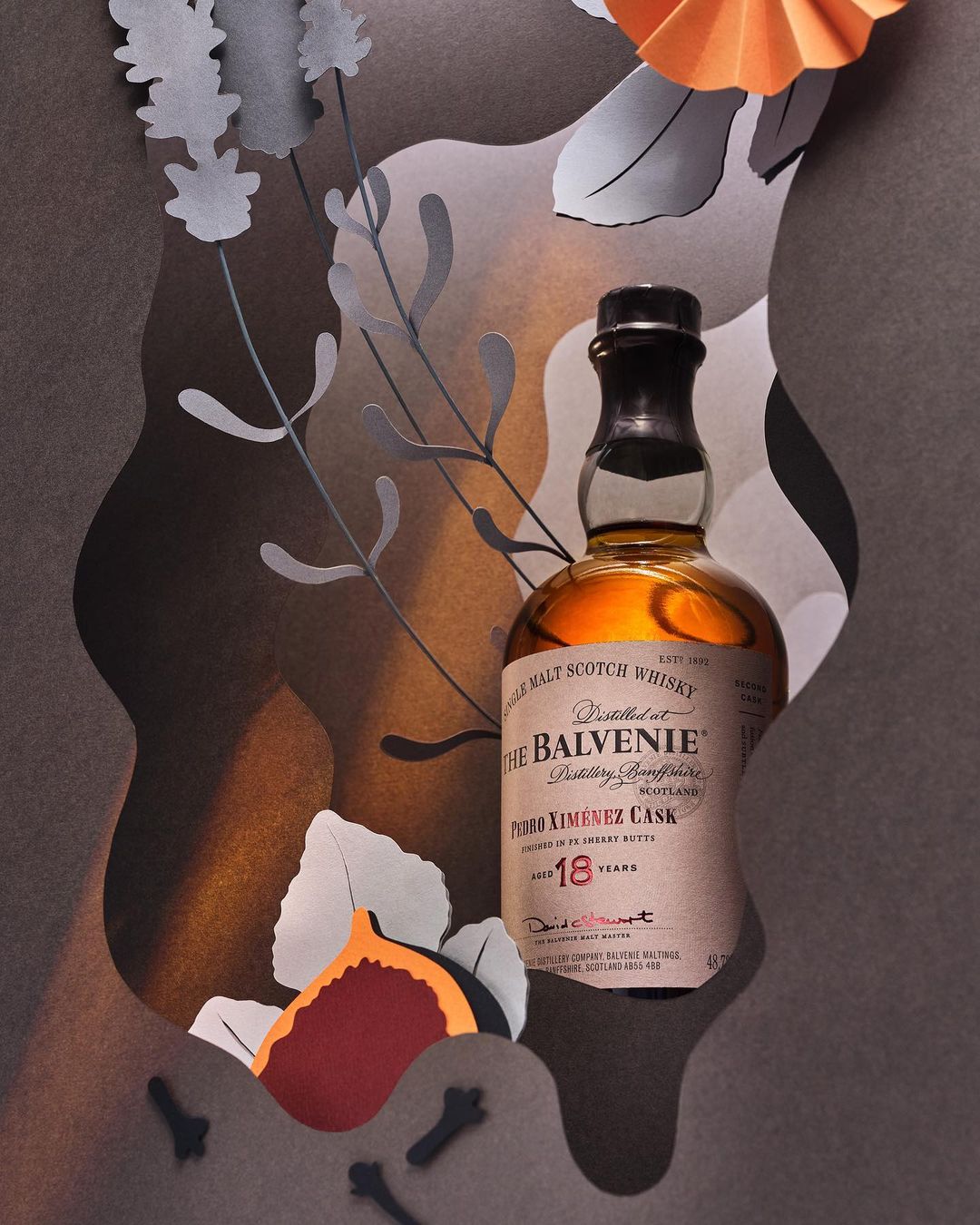 Bottle of The Balvenie single malt Scotch whiskey amidst paper art foliage, featuring an orange fan and brown abstract shapes.