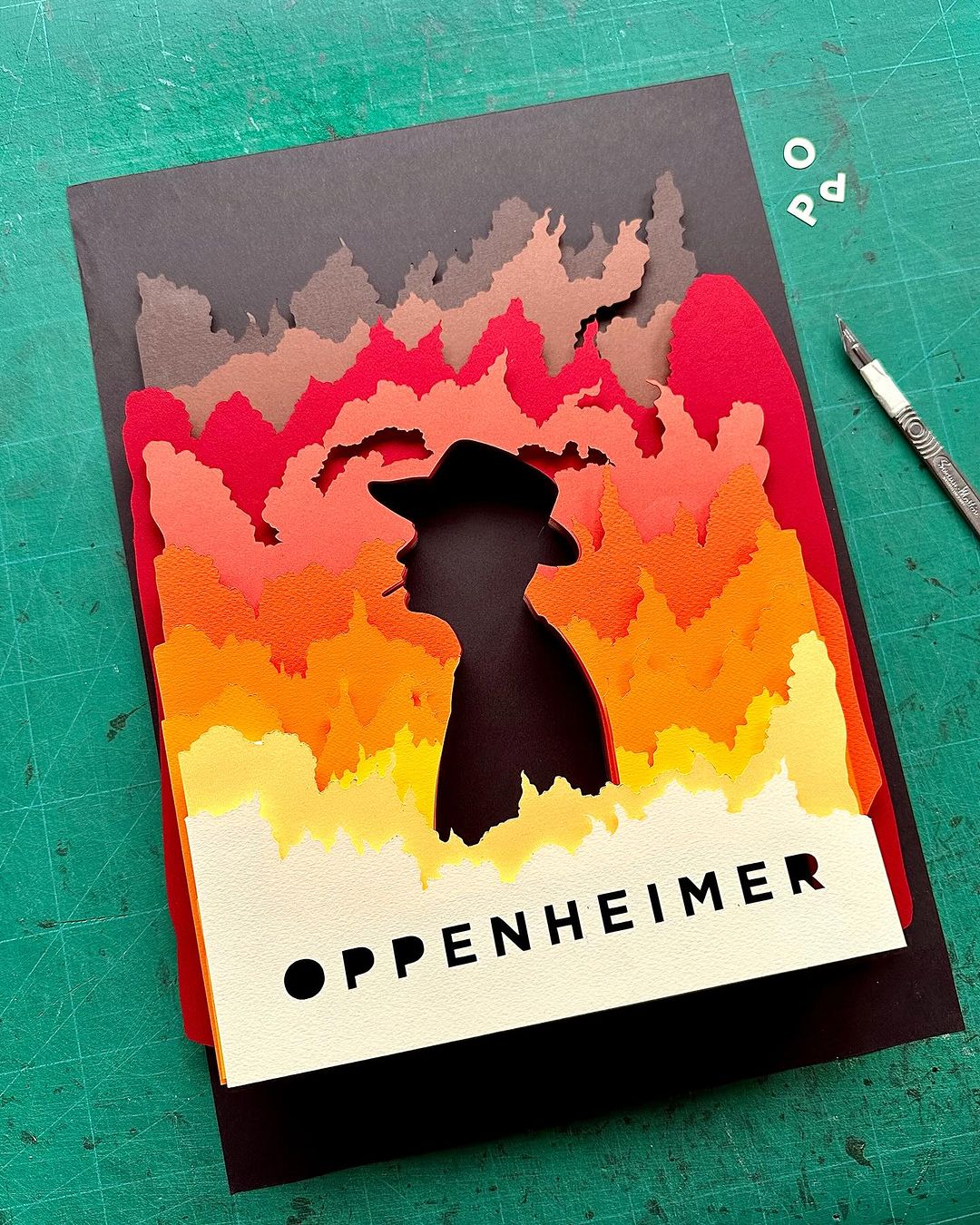 Oppenheimer poster created with paper art technique features a silhouette of a man in a hat against a fiery background.