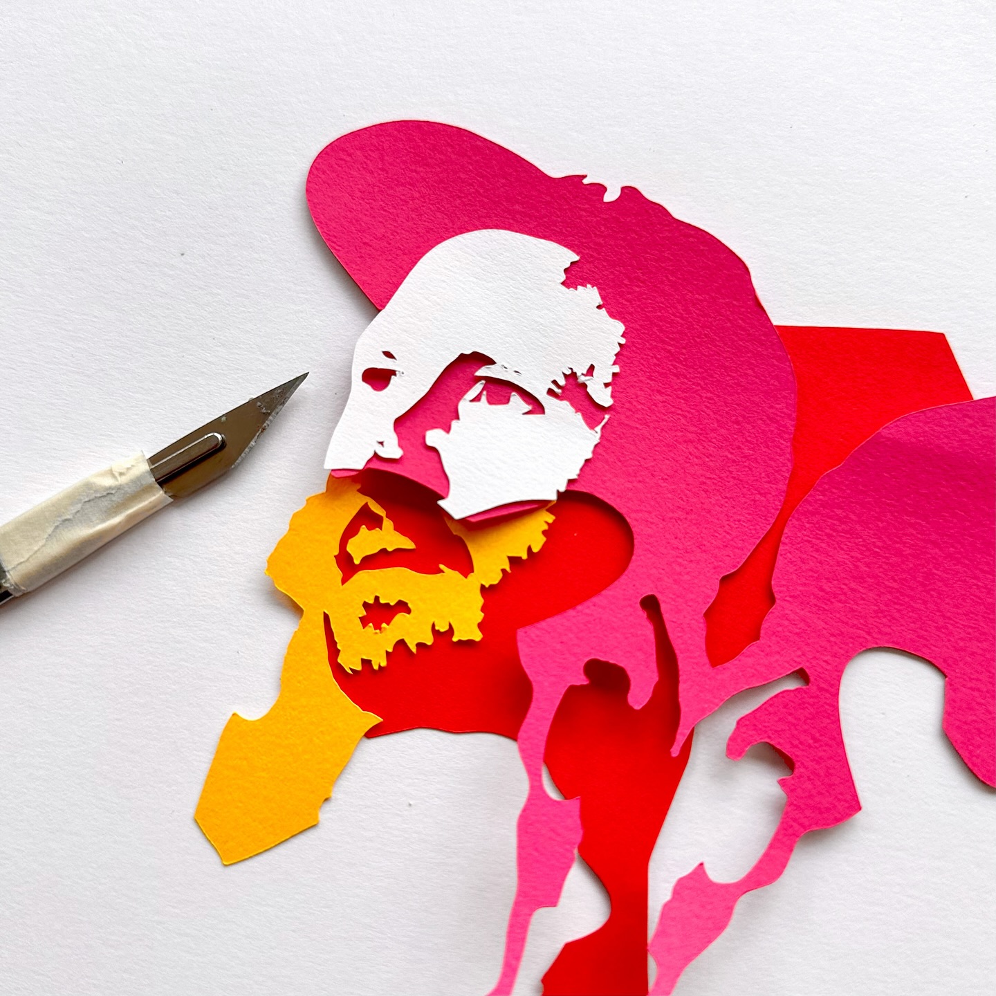 Paper art portrait of a bearded man. The layered paper in shades of white, yellow, red, and pink depicts the man's face, profile, and beard.
