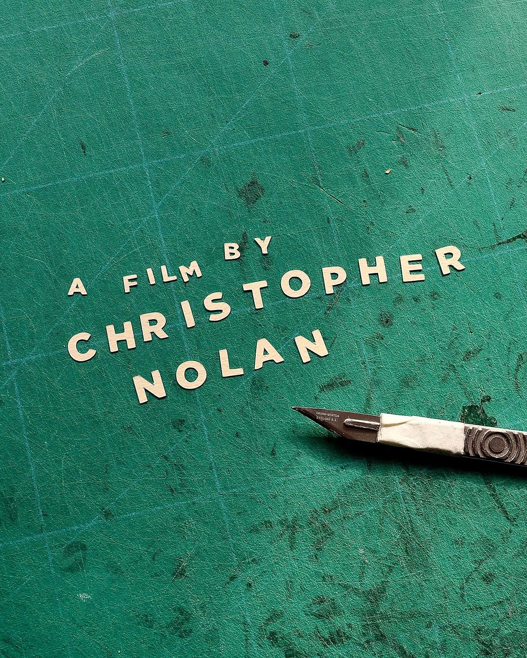 Craft still life of paper cut-out letters spelling 'A Film By Christopher Nolan' with craft knife on green self-healing cutting mat.