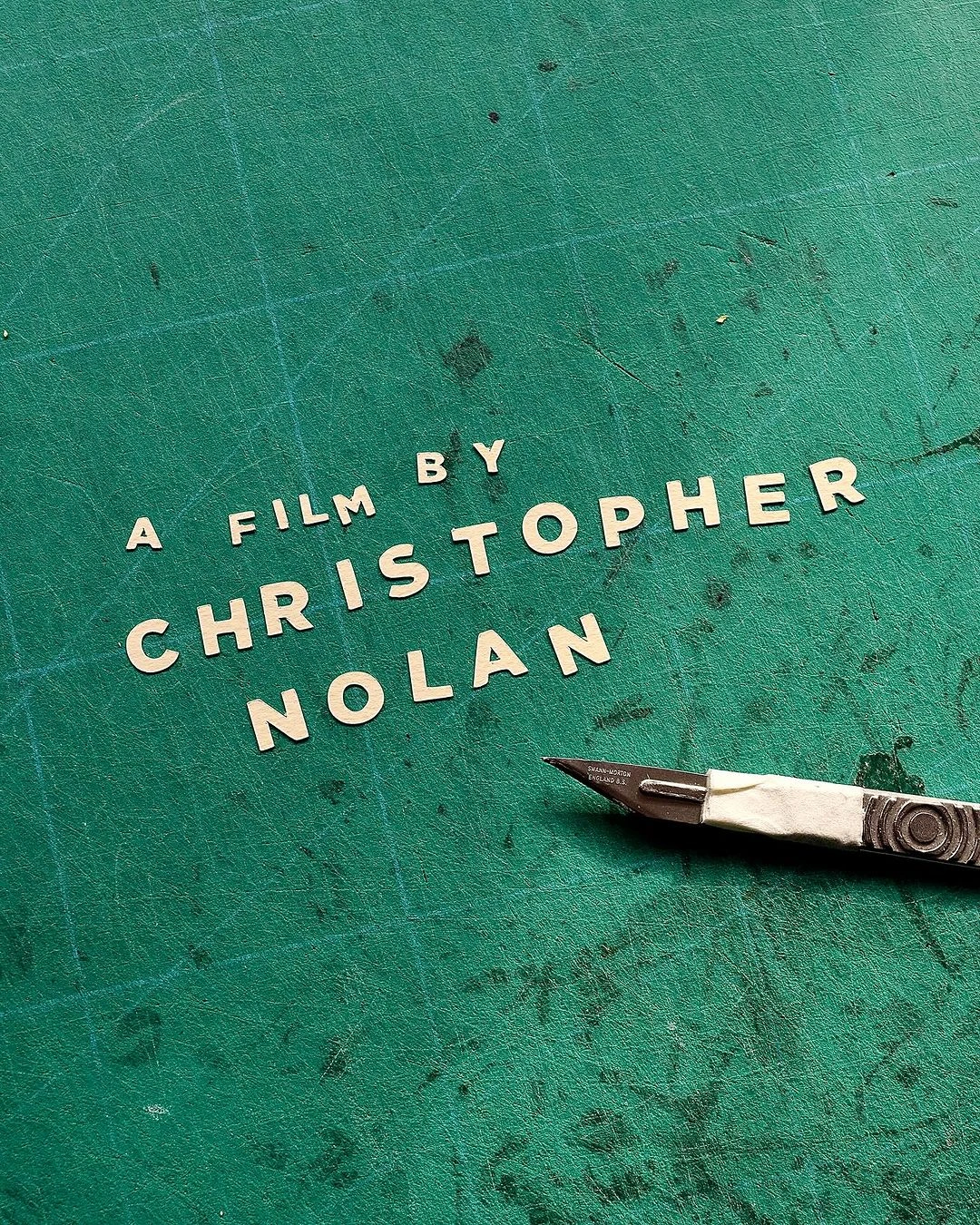 Handcrafted paper lettering that reads A Film By Christopher Nolan arranged on a green cutting mat with a craft knife
