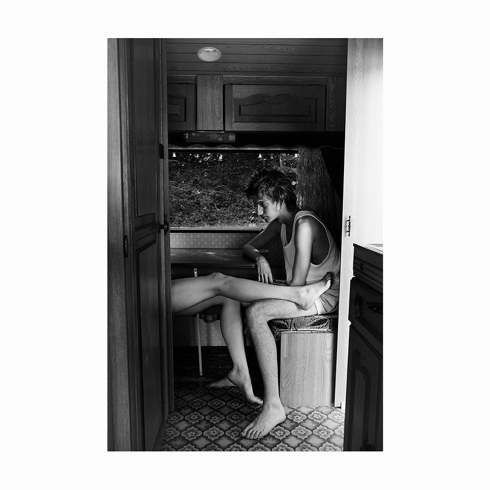 Black-and-white photograph of two people in a camper, one seated with the other's legs resting on their lap, captured in a moment of quiet intimacy.