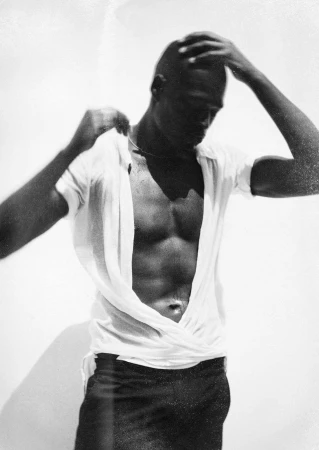 Black man adjusting his unbuttoned white shirt against a plain background in a grainy black and white portrait.