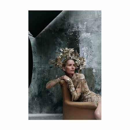 Woman wearing elaborate gold bird headdress and beaded dress, seated in tan armchair against mottled grey wall