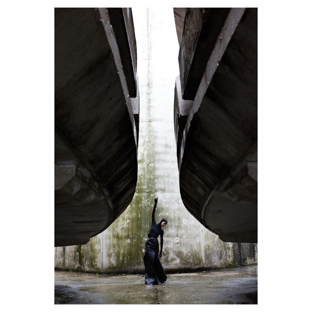 A woman in dark clothing stands between two concrete structures in an outdoor setting, with her arm raised to the sky.