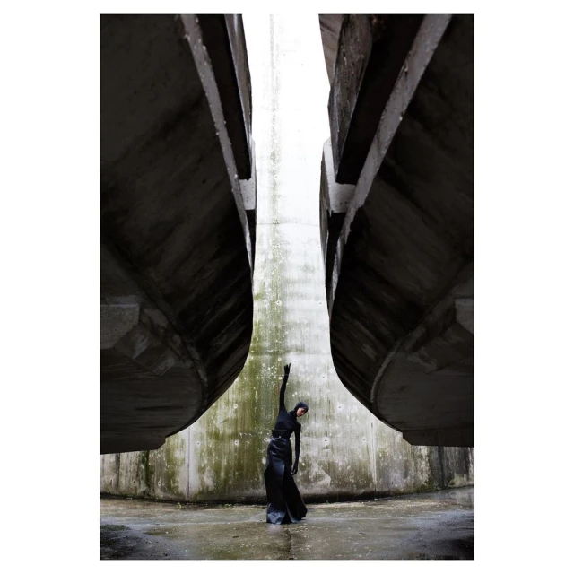 A woman in dark clothing stands between two concrete structures in an outdoor setting, with her arm raised to the sky.
