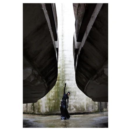 A woman in dark clothing stands between two concrete structures in an outdoor setting, with her arm raised to the sky.