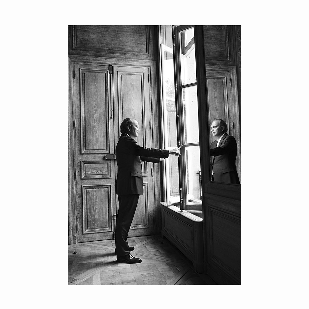 Black and white shot of a man in a suit standing in front of a window and its reflection inside a room with wooden paneling.