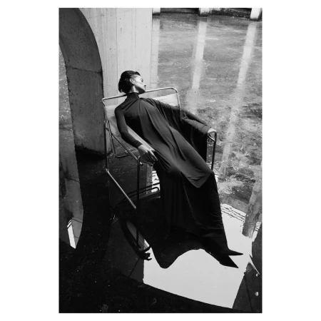 Model in black high neck dress reclining on tubular steel chair over water by P.a Hue De Fontenay