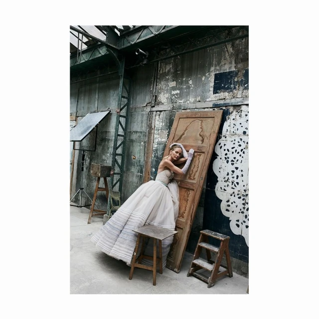 A woman in a strapless gown poses against a weathered door in an industrial space, evoking a vintage fashion editorial.