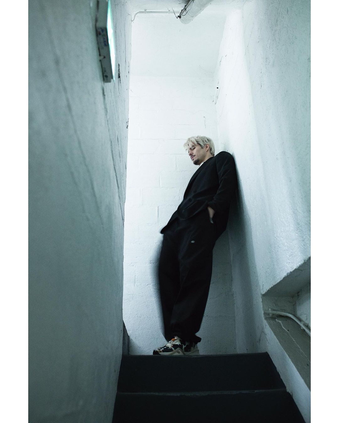 Man in black suit leans against white concrete wall on stairs, indoor shot with diffused light, looking slightly downwards in urban style.