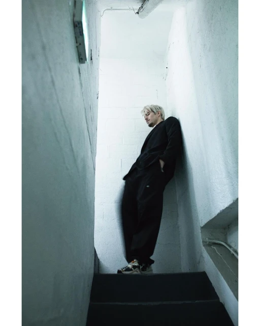 Man in black suit leans against white concrete wall on stairs, indoor shot with diffused light, looking slightly downwards in urban style.