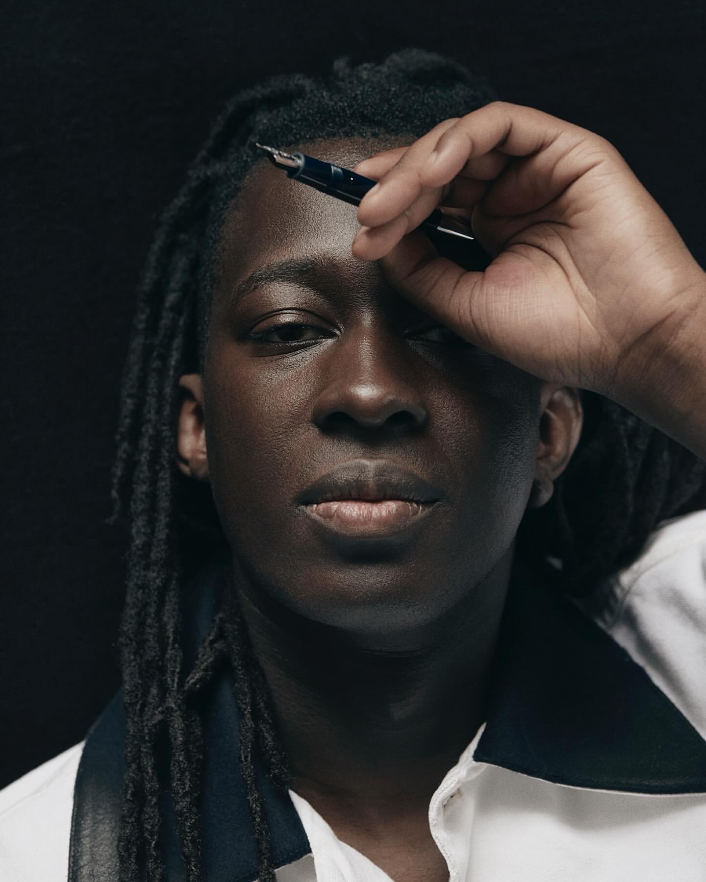 Portrait of a young adult with dark skin and dreadlocks holding a black fountain pen to his forehead against a black background.
