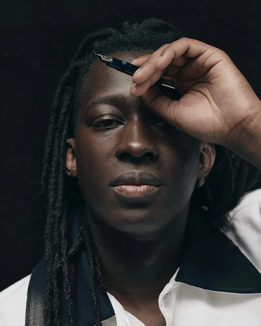 Portrait of a young adult with dark skin and dreadlocks holding a black fountain pen to his forehead against a black background.
