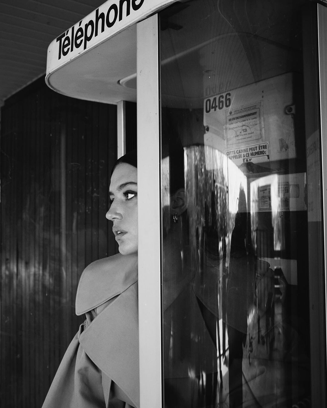 Black and white portrait of a woman inside a telephone booth, wearing a trench coat and looking left with a serious expression.