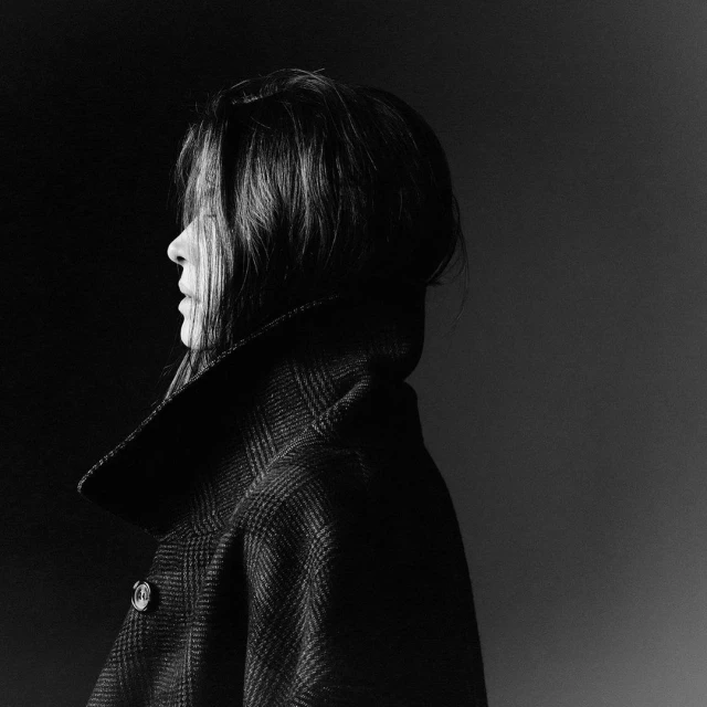 Monochrome portrait of a woman in profile, wearing a patterned wool coat, captured in a dark, minimalist studio setting with diffused lighting.