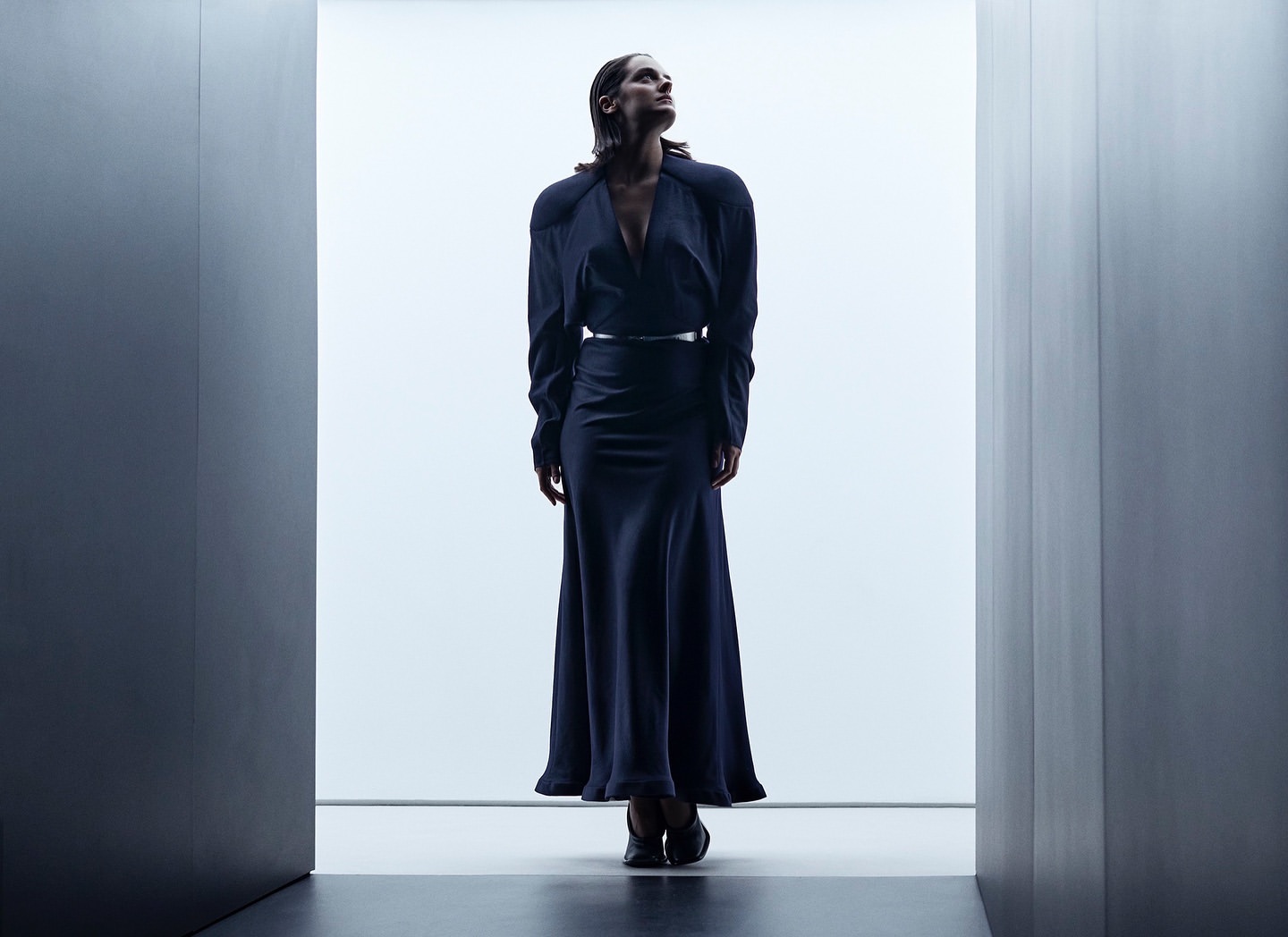 Woman in dark long-sleeve dress standing framed between architectural walls in a minimalist interior shot by P.a Hue De Fontenay