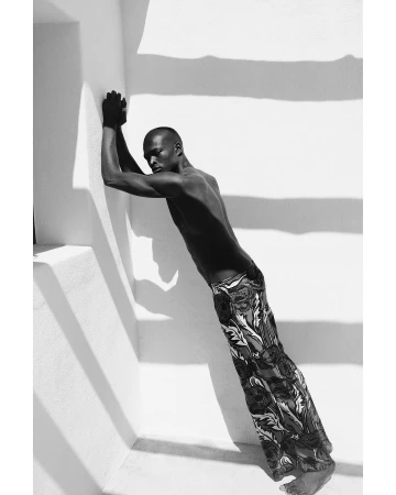 Black and white portrait of a shirtless man leaning against a sunlit white wall wearing patterned trousers by P.a Hue De Fontenay