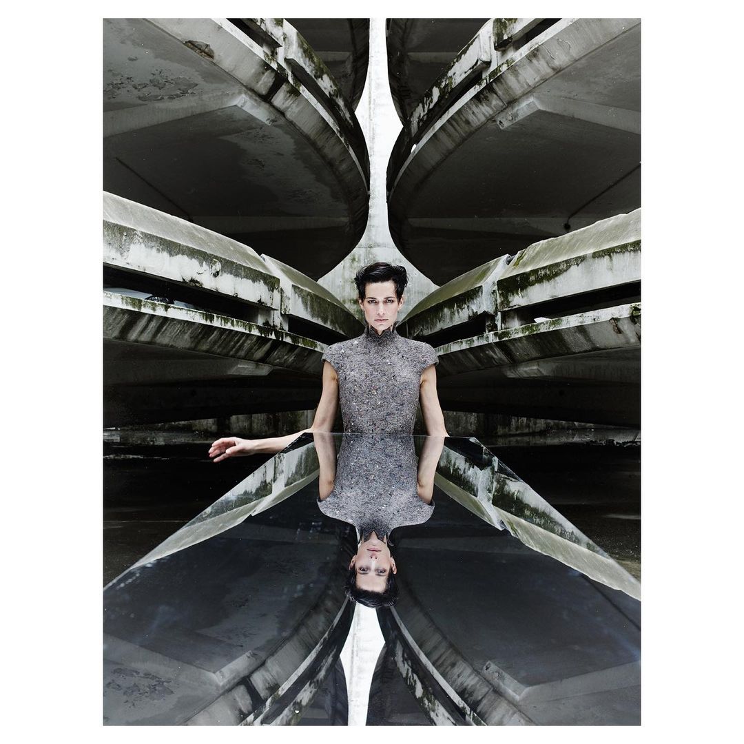 Fashion shot of a woman in a grey textured dress standing in an architectural space with reflective ground mirroring the concrete structures above.