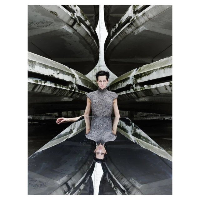 Fashion shot of a woman in a grey textured dress standing in an architectural space with reflective ground mirroring the concrete structures above.