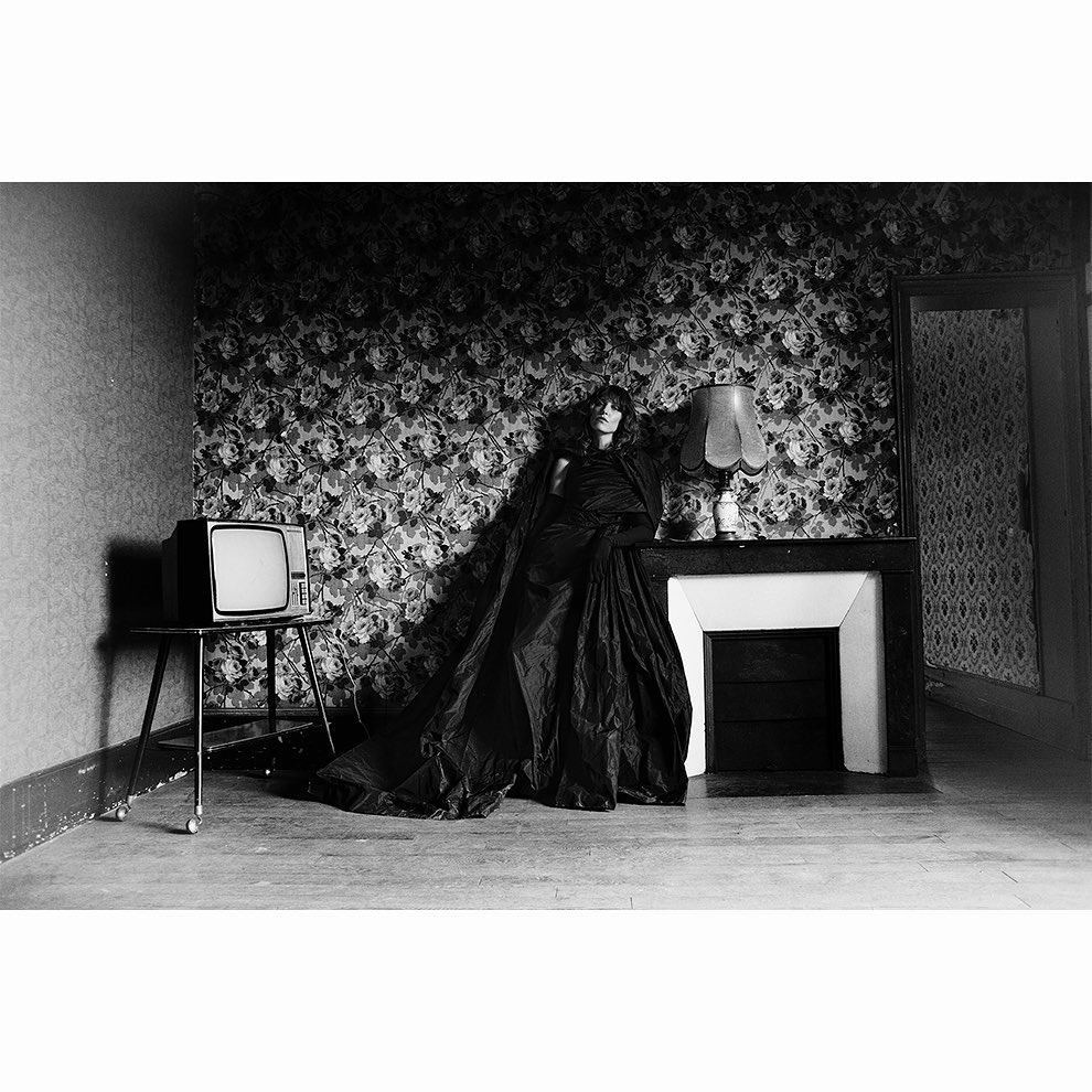 Monochrome portrait of a woman in a dark gown leaning against a fireplace, beside a vintage television in an ornate interior room.