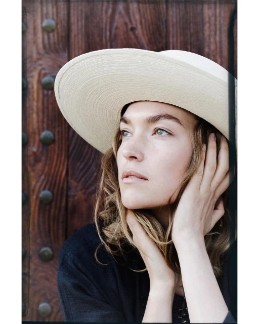 Woman in a wide-brimmed straw hat poses in front of a weathered wood door with metal accents in a close-up portrait shot.
