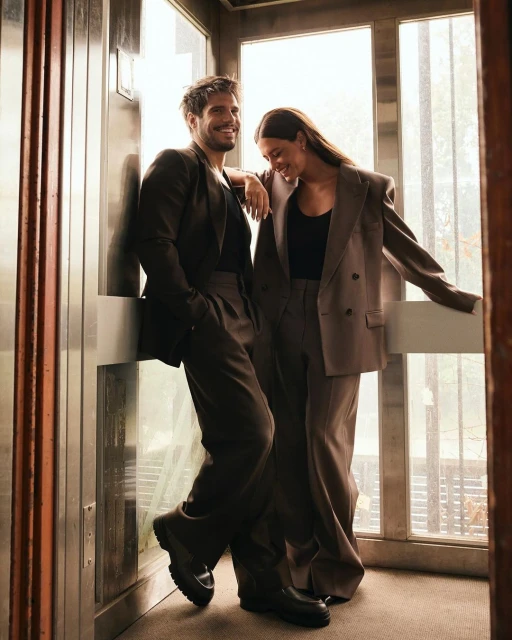 A smiling man and woman in fashionable suits pose in an elevator, creating a stylish portrait with natural lighting and a neutral color palette.