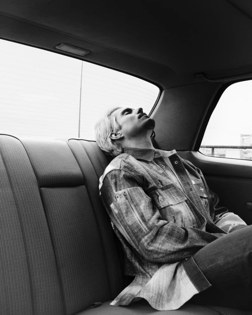 Black and white portrait of a male passenger with blonde hair reclining in the backseat of a vintage vehicle looking out the window.