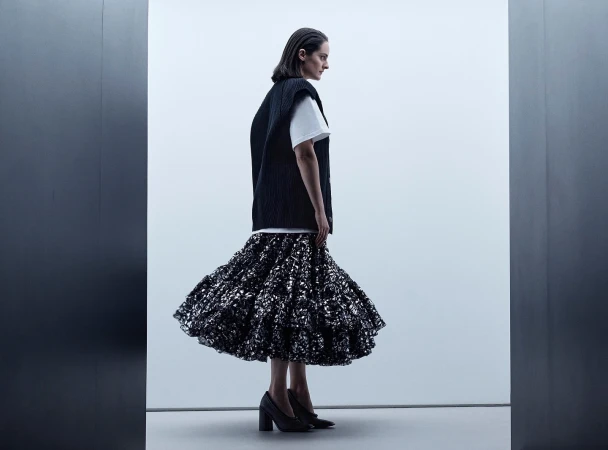 A woman wearing a white t-shirt, black vest, and dark ruffled skirt stands against a bright studio background.