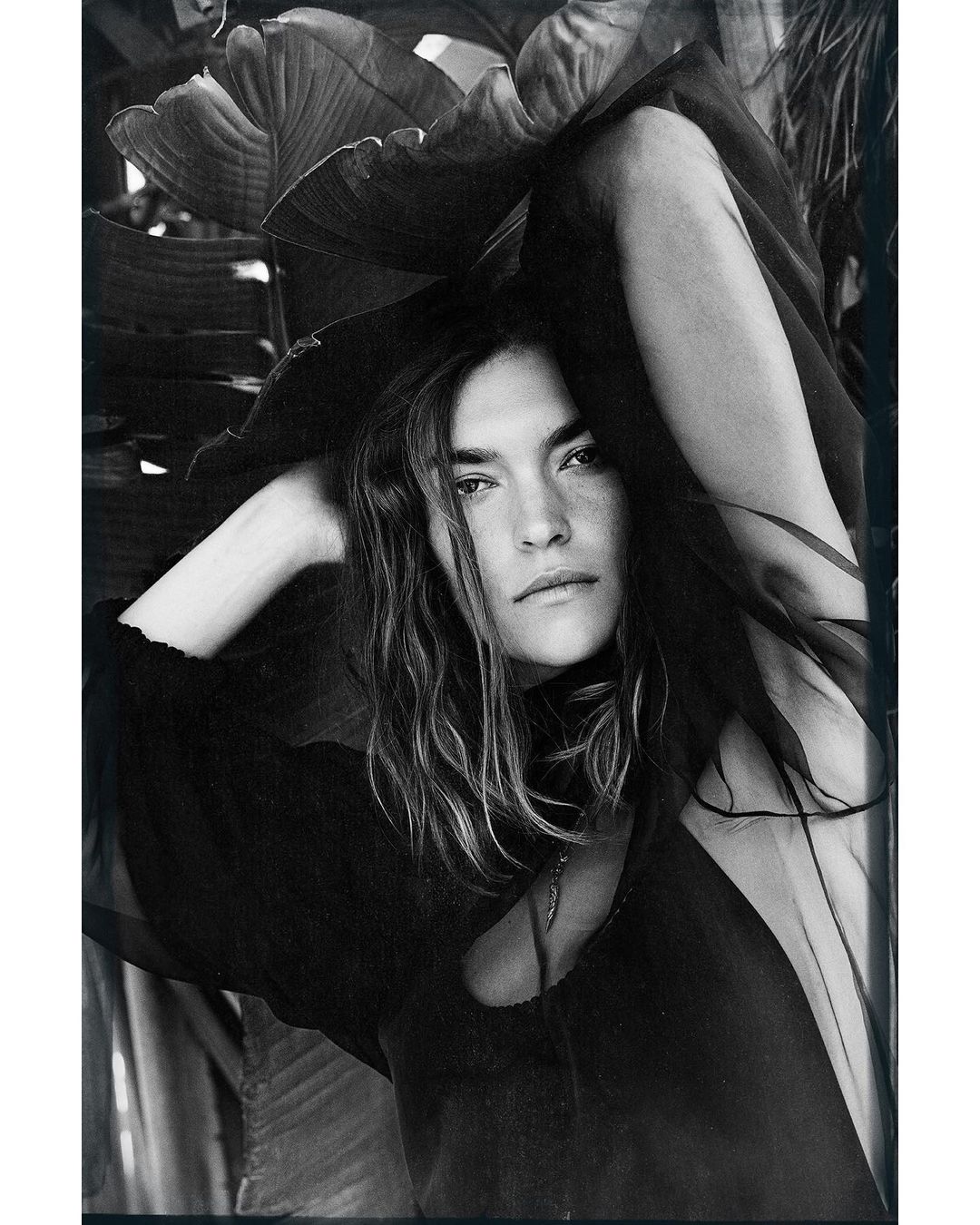 Monochrome portrait of a woman with long hair posing outdoors with arms raised, framed by large tropical leaves. Fashion editorial.