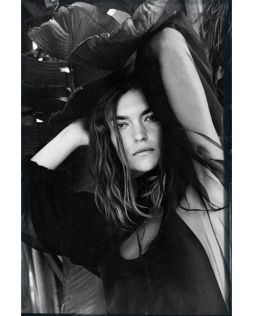 Monochrome portrait of a woman with long hair posing outdoors with arms raised, framed by large tropical leaves. Fashion editorial.