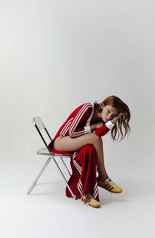 A redhead woman in a red Adidas jumpsuit poses in a studio sitting on a clear chair. She wears yellow sneakers and has a pensive expression.