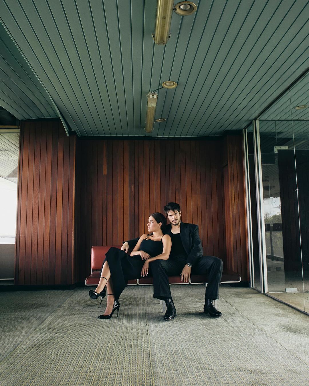 A stylish couple in black attire poses on a bench in an indoor space featuring wood paneling and large windows, capturing a modern, elegant moment.