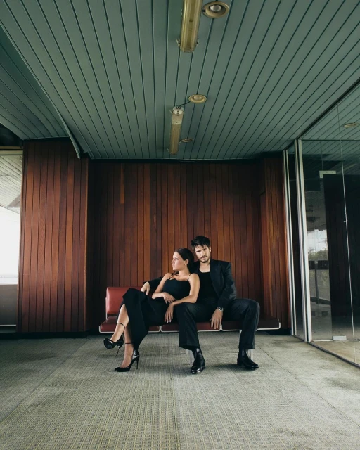 A stylish couple in black attire poses on a bench in an indoor space featuring wood paneling and large windows, capturing a modern, elegant moment.