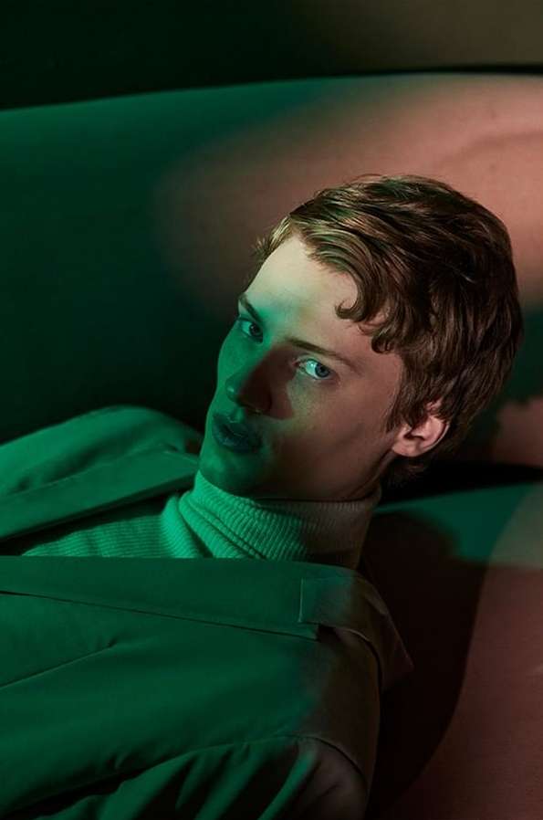 Portrait of a young adult male with brown hair, wearing a cream-colored turtleneck and sage green jacket, laying down with green lighting.