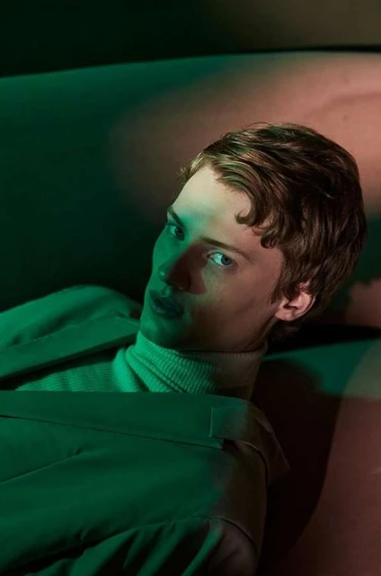 Portrait of a young adult male with brown hair, wearing a cream-colored turtleneck and sage green jacket, laying down with green lighting.