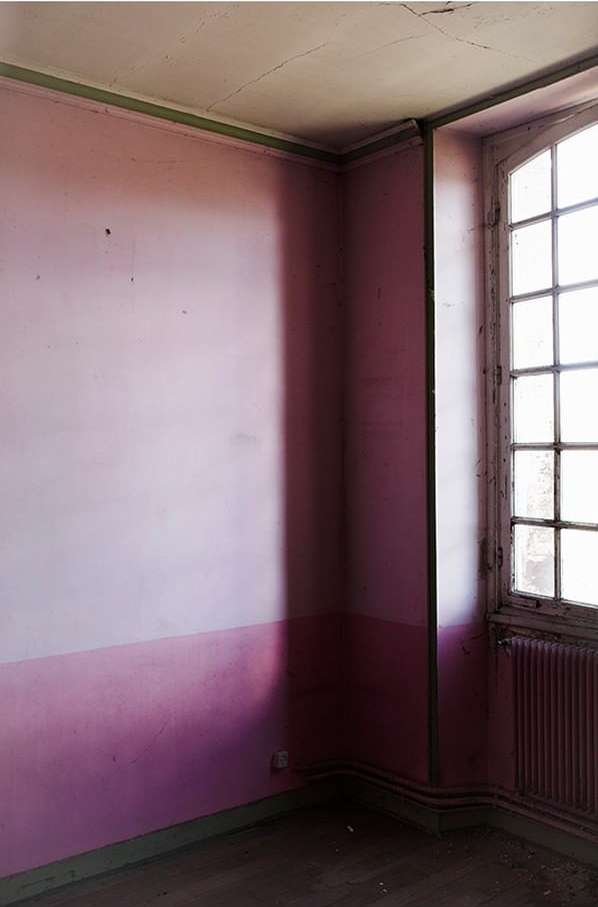 Dilapidated pink room interior featuring a window with natural light, faded walls, and minimal furniture, evoking a sense of decay.