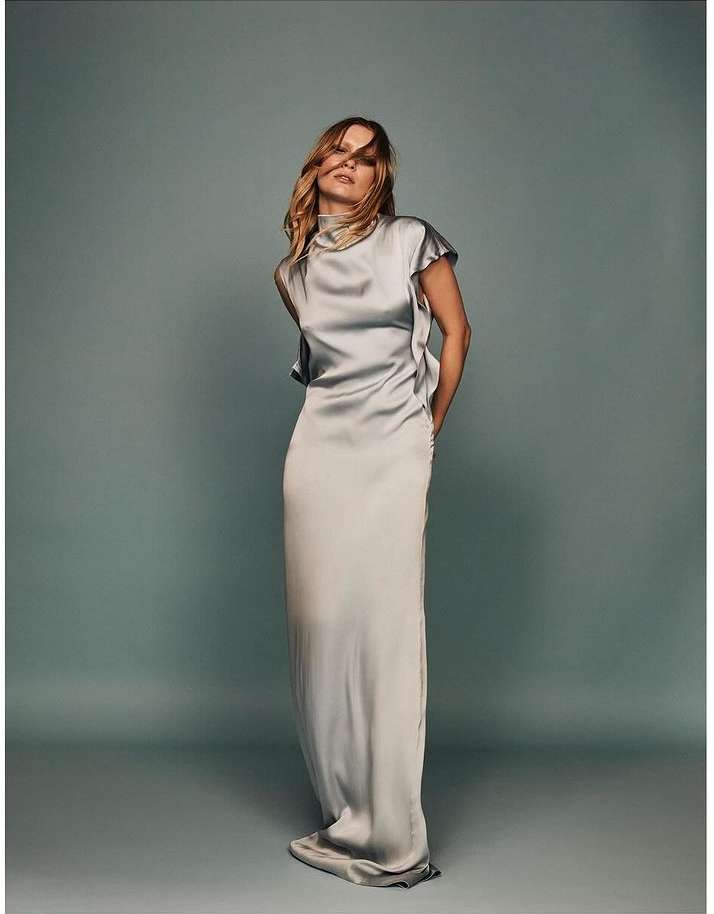 Elegant woman in a light gray, floor-length satin dress with a high neckline, posing in a studio with a light gray backdrop. Fashion photography.