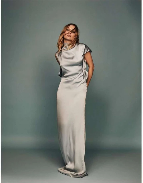 Elegant woman in a light gray, floor-length satin dress with a high neckline, posing in a studio with a light gray backdrop. Fashion photography.