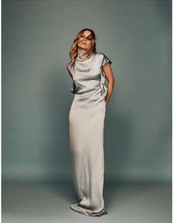 Elegant woman in a light gray, floor-length satin dress with a high neckline, posing in a studio with a light gray backdrop. Fashion photography.