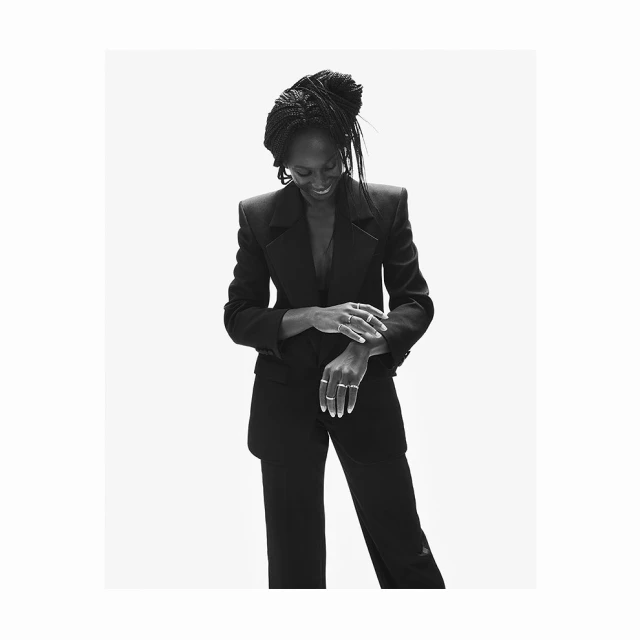 Black and white portrait of a Black woman in a stylish black suit, looking down with a slight smile, standing in a bright photo studio.