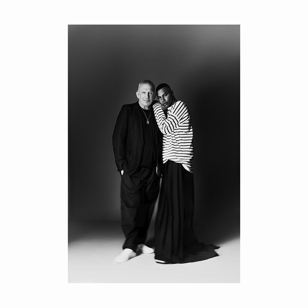 Black and white portrait of two men: one in a suit, the other in a striped shirt and long skirt, standing in a photo studio.