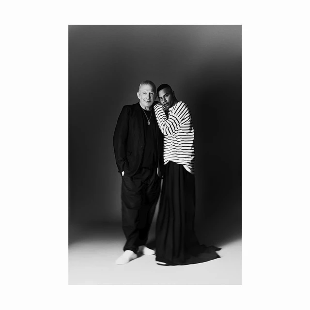 Black and white portrait of two men: one in a suit, the other in a striped shirt and long skirt, standing in a photo studio.